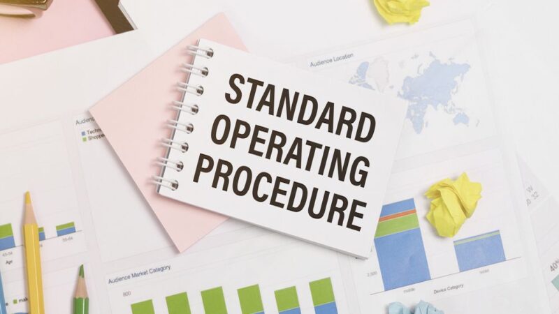 Standard Operating Procedure text on paper in open diary with spectacles, colourful push pin, pen and calculator on the wooden table - business and finance concept
